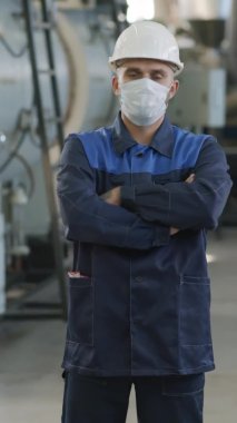  Üniformalı erkek fabrika işçisinin dikey portresi, kollarını kavuşturan ve iş yerinde kameraya poz veren kasklı ve yüz maskesi.