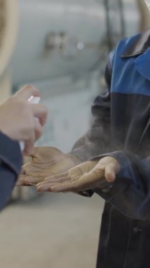  Yüz maskesi takmış siyah erkek bitki işçisinin avuç içlerine el dezenfektanı sıkan tanınmayan bir yöneticinin dikey yakın çekimi fabrikadaki vardiyasına başlıyor.