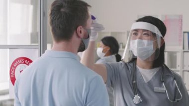Koruyucu yüz kalkanı takan ve hastaların sıcaklığını temassız termometreyle ölçen hemşirenin yavaşlığıyla dikiz görüş ortamına yaklaş