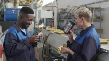  Medium shot of cheerful male and female factory workers in uniforms laughing and chatting while enjoying their sandwiches during lunch break in workplace