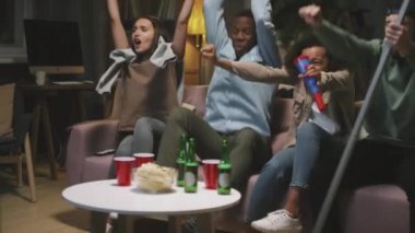 Ağır çekimde, çok ırklı arkadaş grubunun TV 'de birlikte maç izlediği, kanepeye oturduğu, abur cubur ve bira içtiği, hedefi kutladığı, birbirine sarılıp beşlik çaktığı bir fotoğraf.