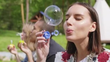 Kamp alanında sabun köpüğü üfleyen genç bir kadının portresi. Arka planda oturan arkadaşlarıyla kameraya bakıyor. Hepsi iyi vakit geçiriyor.