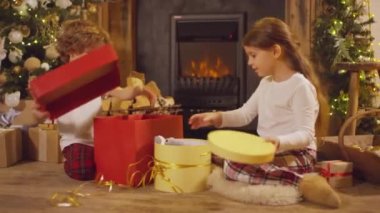 Önünde oturan iki genç çocuk içi doldurulmuş oyuncak ve ahşap uçakla Noel hediyesi kutularını açıyor.