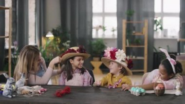 Paskalya atölyesinde çiçeklerle süslenmiş saman şapka deneyen ve neşeli kız ve oğlanlarla konuşan mutlu bir genç kadın resmi.