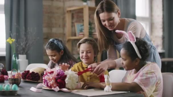 中等比例的年轻女子微笑着 帮助三个孩子坐在桌旁 在复活节用鲜花和缎带装饰草帽 — 图库视频影像