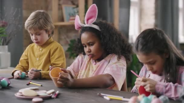 Közepes lövés aranyos gyerekek ül asztalnál a művészeti és kézműves osztály és díszítő húsvéti tojás és ceruza csésze