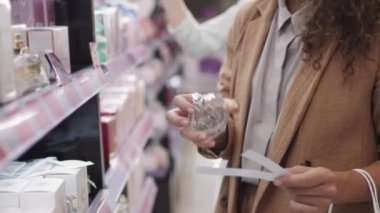 Orta boy, uzun kıvırcık saçlı, güzel ve çekici bir bayanla yakın çekim. Güzellik salonunda duruyor. Yeni bir koku seçiyor, üzerine parfüm sıkıyor ve kokluyor.