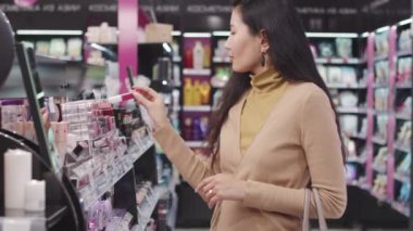 Kozmetik mağazasında ruj seçen çekici Asyalı kadının yavaş çekimleri. Renklere bakarak test uygulaması.