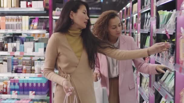 化粧品店で美容製品の買い物袋を持つ2つのエレガントな多民族の女性のスローモーション中ショット 一緒に顔シートマスクを選択 — ストック動画