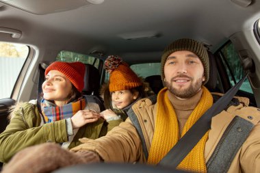 Sıcak kış giysileri içinde, karısının ve kızının karşısında direksiyon sallarken arabanın ön camından bakan mutlu ve aktif genç bir adam.