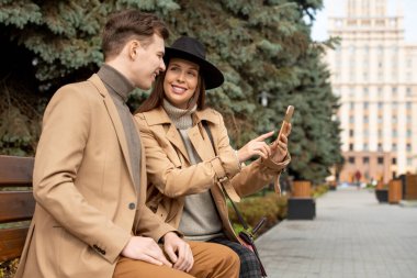 Mutlu, genç, sevgi dolu bir çift şık giyimli, kozalaklı ağaçların yanında oturmuş, akıllı telefonunun ekranına gülümseyen kızı işaret ediyor.