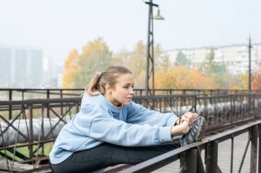 Köprüde duran genç aktif sporcu kadın kollarını gerdi ve sağ bacağını metalik çitlere dayadı ve sabahları dışarıda egzersiz yapıyor.
