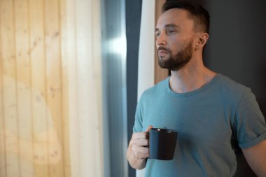 Gri tişörtlü genç düşünceli ve rahat bir adam elinde fincanla sıcak çay ya da kahve tutuyor ve evde otururken büyük pencereden dışarı bakıyor.
