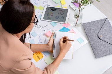 Genç esmer iş kadını ana hedefleri ve çalışma noktaları olan büyük bir belgenin üzerine eğilerek defter sayfasına notlar alıyor.