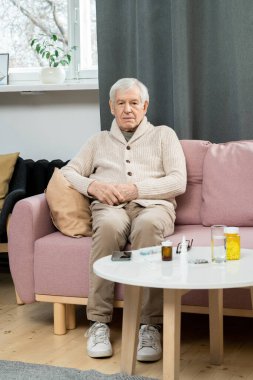 Günlük kıyafet giyen hasta kıdemli bir adam küçük bir masada oturuyor. Yanında ilaçlar ve bir bardak su var. Oturma odasında doktor bekliyor.