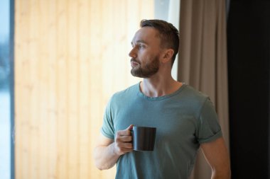 Genç sakallı, rahat, gri tişörtlü, elinde fincanla elinde sıcak çay ya da kahveyle evde otururken büyük pencereden içeri bakan adam.