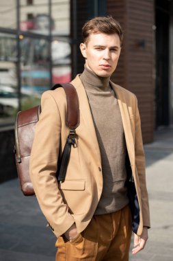 Bej ceketli ve gri kazaklı genç ciddi işadamı kameranın önünde modern binaya karşı dikilirken yana bakıyor.