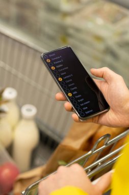 Akıllı telefonlu genç erkek kurye marketten taze ekmek ve sütle alışveriş arabasının üzerinden almak için listeyi karıştırıyor.