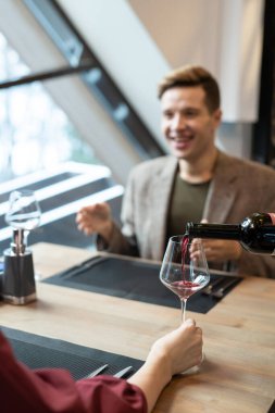 Genç bir kadının eli, mutlu erkek arkadaşının önünde masaya oturmuş elinde şarap kadehi tutarken üstüne kırmızı şarap koyuyor.