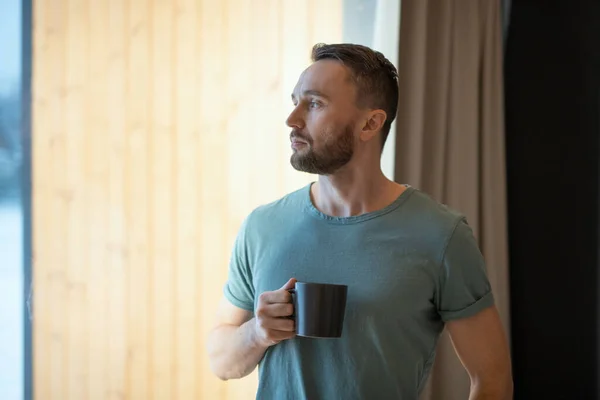 若いですBeadedリラックスした男でグレーTシャツ保持マグカップとともに熱いお茶やコーヒーと家にいながら大きな窓を通して見る — ストック写真