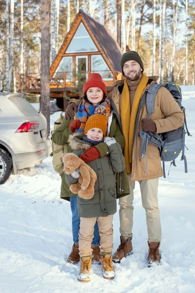 穿着温暖冬衣的三口之家的年轻人站在摄像机前 面对着他们的乡间别墅 看着外面的你 — 图库照片
