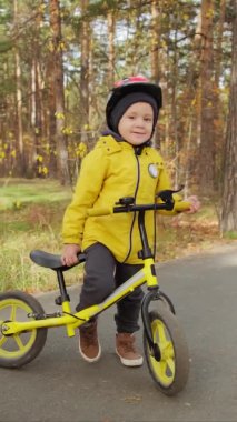 Güneşli bir sonbahar gününde kasklı, dengeli bisikletle parkta poz veren 5 yaşında tatlı bir çocuğun dikey çekimi.