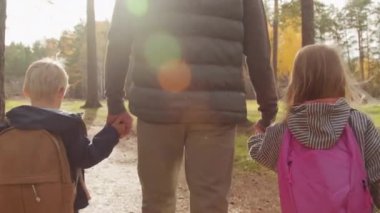 Tanımlanamayan babayı yakından takip edin. Sevimli kız ve oğlanın ellerini tutun ve güneşli bir sonbahar gününde onlarla birlikte parkta veya ormanda yürüyün.