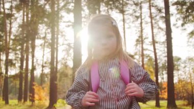 Güneşli bir sonbahar gününde, sırt çantasıyla parkta kameraya poz veren 5 yaşındaki tatlı bir kızın portre fotoğrafı.