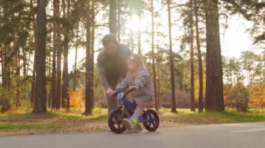 Güneşli bir sonbahar gününde, babasının 5 yaşındaki küçük kızının asfalt yolda dengeli bisiklet sürmesine yardım ederken çekilmiş bir fotoğrafı.
