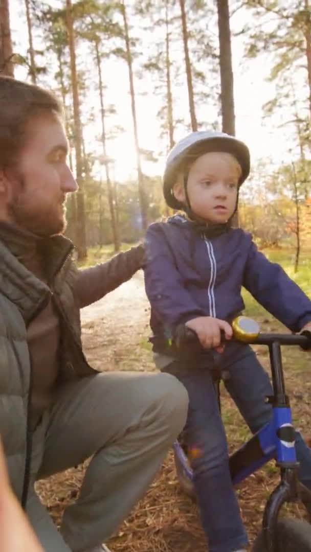 垂直のハメ撮りの父保持カメラと撮影自分自身と彼の小さな息子オンバランス自転車で公園で晴れた秋の日 — ストック動画