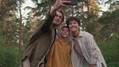 Yeşil yaz ormanlarında aile yürüyüşü sırasında akıllı telefonlu neşeli bir adamın karısıyla ve oğluyla selfie çekerken orta açı çekimi.