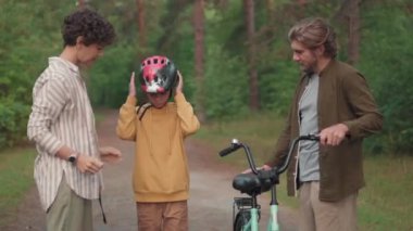 Üç çocuklu, mutlu, beyaz bir ailenin yaz gününü ormanda geçirdiği orta boy bir fotoğraf. 12 yaşında bir çocuk bisiklete biniyor ve ailesi izliyor.