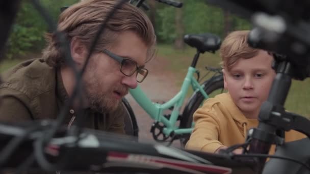 中程度のクローズアップで 彼のお父さんを説明する若い男の子の遅い自転車を修正する方法 家族の散歩中に森の中で屋外で不法占拠 — ストック動画