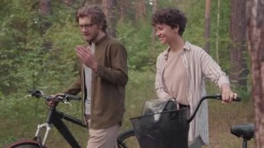 Romantik bir buluşmada genç beyaz bir çiftin, ormanda bisikletle dolaşırken ve sohbet ederken orta ölçekli bir fotoğrafı.