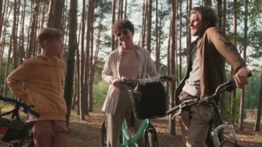 Neşeli beyaz bir ailenin, güneşli bir günde ormanda bisikletle durup sohbet ederken ve kahkahalar atarken orta boy bir fotoğrafı.