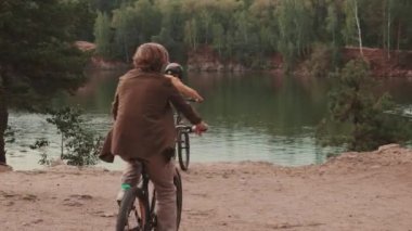 Aktif ebeveynlerin ve 12 yaşındaki çocuklarının parkta bisiklet sürerken, göletin önünde durup doğanın tadını çıkarırken tam bir dikiz görüntüsü.