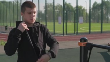 Yazın spor sahasına gelen, spor çantalı ve eşofmanlı genç beyaz adamın spor barında spor yapmadan önce ısınmaya başladığı orta boy fotoğrafı.