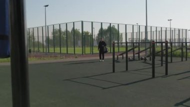 Siyah eşofmanlı, spor çantalı beyaz bir adamın yaz günü spor salonuna gelip antrenmana başlamadan önce üst ceketini çıkarmasıyla dolu bir fotoğraf.