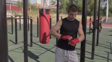 Beyaz tenli genç bir sporcunun orta boy portresi ellerini kırmızı bir bandajla sarmalayıp spor sahasında kum torbasının yanında duran kameraya bakıyor.