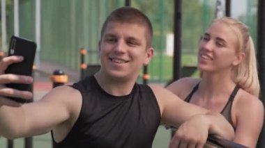 Açık hava antrenmanı sırasında kameraya gülümseyen akıllı telefondan selfie çeken genç ve neşeli bir çiftin orta boy fotoğrafı.