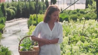 Kafkas kadın bahçıvanın yağmurdan sonra güzel yeşil bahçe boyunca hasır sepetle kameraya doğru yürüdüğü orta boy bir fotoğraf. Hayranlıkla bitkilere bakıyordu.