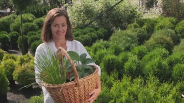 Orta boy PAN portresi, elinde hasır sepet yeşil bitkiler tutan beyaz kadın yaz bahçesinde kameraya bakıyor.