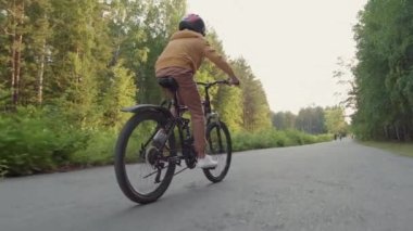 12 yaşında, kasklı bir çocuğun yaz mevsiminde parkta bisiklet sürerken yavaş çekimde dikiz aynasından çekilmesi.