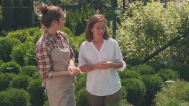 Beyaz bir kadının yaz bahçesine hayran olduğu orta boy bir fotoğraf. Genç bir bahçıvan, bahçede yetişen bitkilerden ve çiçeklerden bahsederken, bahçede gezinti yaparken.