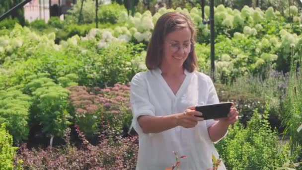 Medium shot of cheerful caucasian woman admiring beautiful green plants and flowers in summer yard under sunlight, taking photos of them on her smartphone
