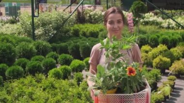 Orta boy PAN portresi. Bahçıvan olarak çalışan gülümseyen bir kız portresi. Saksı bitkileri ve çiçeklerle dolu bir kutuya gülümsüyor.