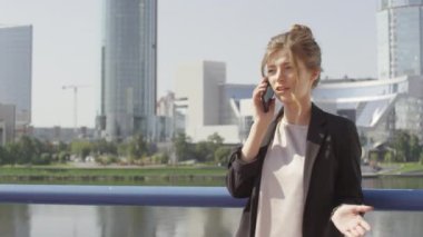 Güneşli bir günde şehir merkezinde köprü üzerinde cep telefonuyla konuşan çekici genç iş kadınının orta boy fotoğrafı. Arka planda modern binalar ve nehir