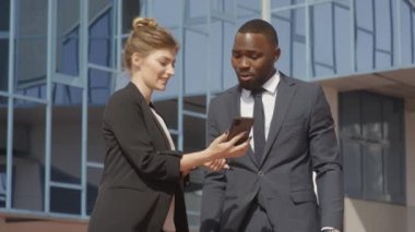 Çok ırklı iş çiftlerinin yavaş çekimleri akıllı telefon ekranlarına bakıp güneşli bir günde modern ofis binasının yanında duran bir şeyi tartışıyorlar.