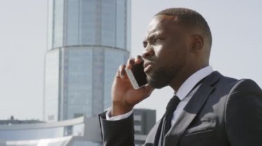 Modern iş bölgesinde arka planda uzun ve modern ofis binası olan akıllı telefondan konuşan ciddi Afro-Amerikan iş adamının düşük açılı görüntüsü.