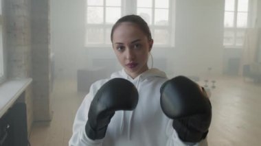 Çatı katındaki boks eğitimi sırasında kameraya bakarak siyah boks eldiveni giyen çekici genç bir kadının yavaş çekimde orta boy portresi.